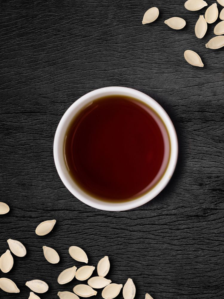 Pumpkin Seed Oil in a small white bowl, surrounded by pumpkin seeds on a dark wooden surface.